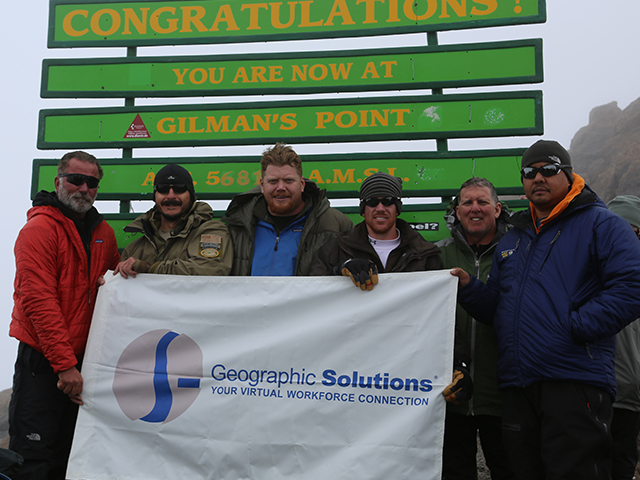 In this picture, a group of employees pose with a sign that bears Geographic Solutions’ logo at Mt. Kilimanjaro.